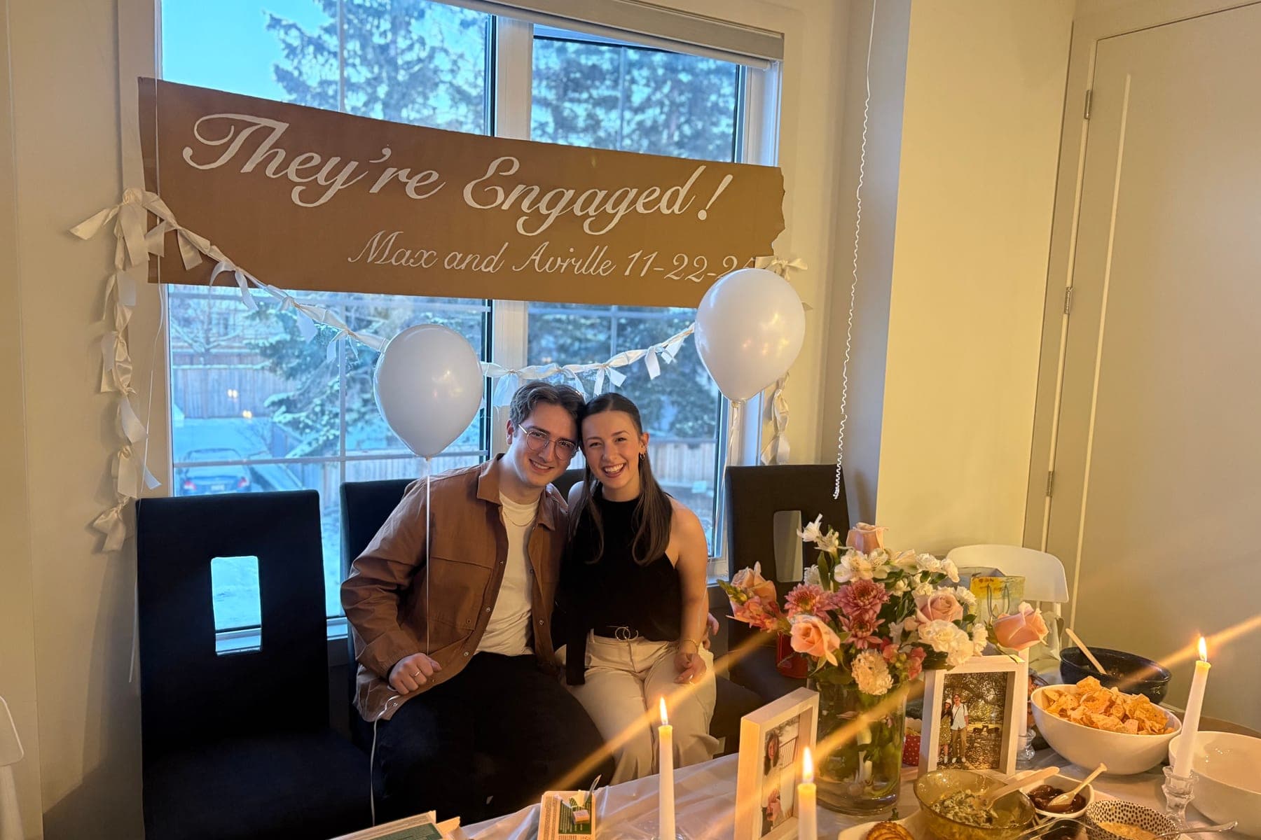 Avrille and Maxwell beneath a hand-lettered “They’re Engaged!” sign.
