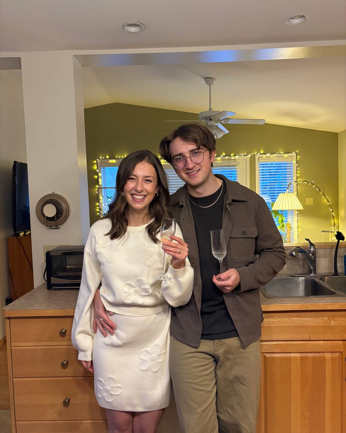 Avrille and Maxwell raising champagne glasses in a kitchen strung with lights.